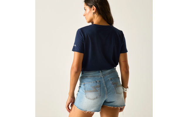 Person von hinten vor hellem Hintergrund, trägt dunkelblaues T‑Shirt und helle Jeans-Shorts mit ausgefranstem Saum, dunkle Haare, Kopf nach links gedreht, Ohrringe und Armbänder sichtbar
