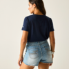 Person von hinten vor hellem Hintergrund, trägt dunkelblaues T‑Shirt und helle Jeans-Shorts mit ausgefranstem Saum, dunkle Haare, Kopf nach links gedreht, Ohrringe und Armbänder sichtbar
