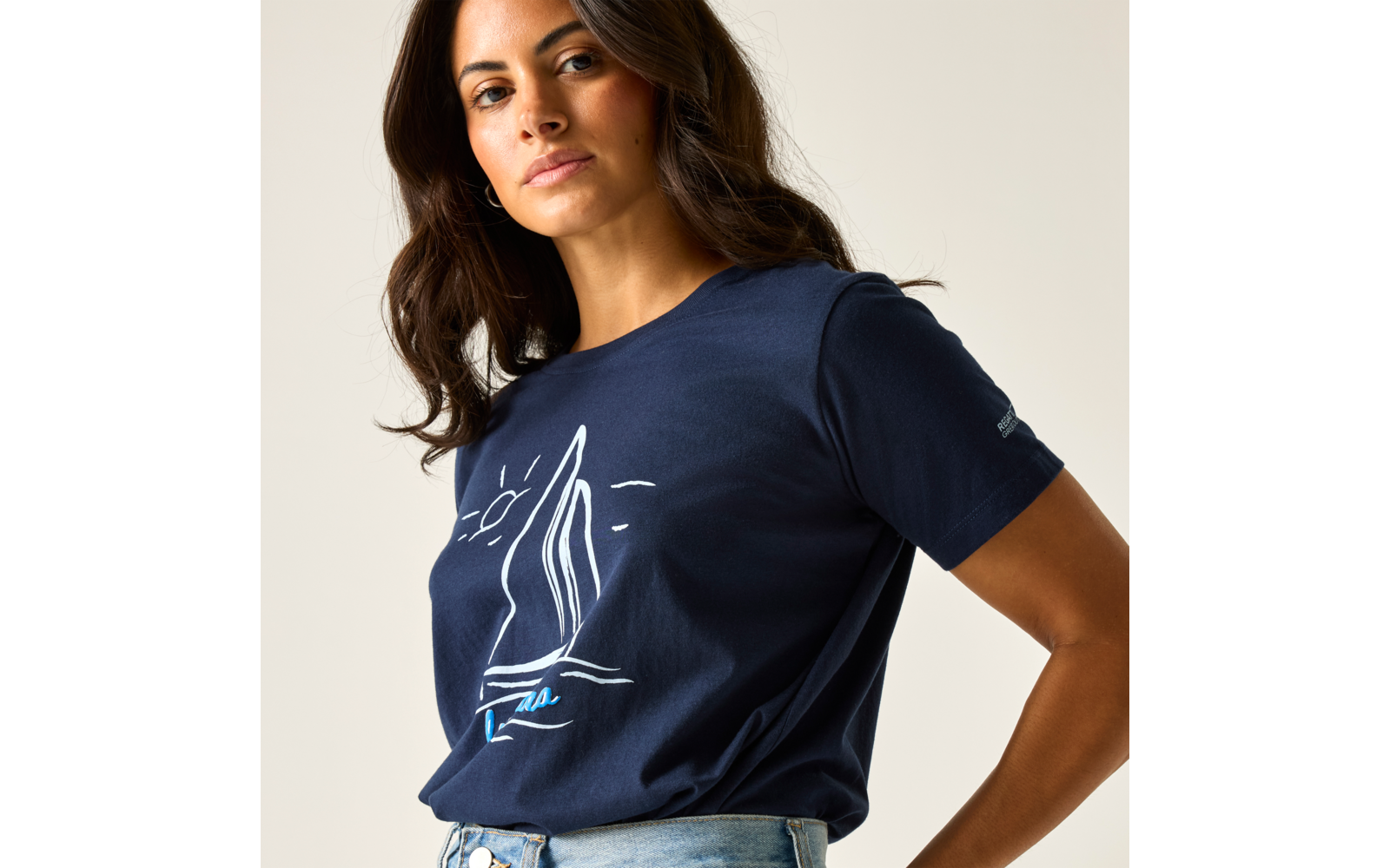 Frau mit schulterlangem dunklem Haar, trägt dunkelblaues T‑Shirt mit weißem Segelboot-Print und helle Jeans