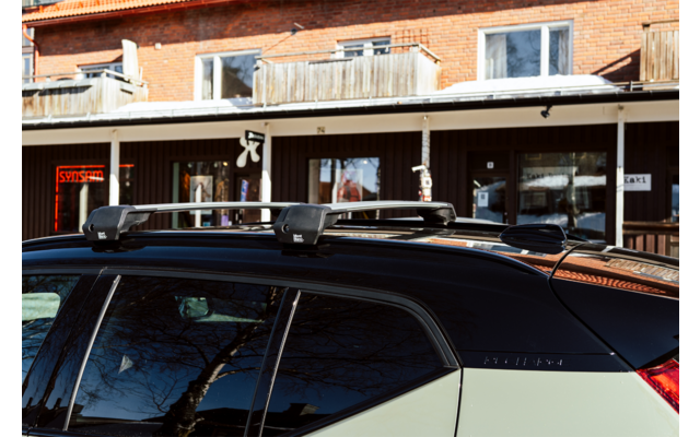 Dach eines hellen Autos mit schwarzem Dach und montierten Querträgern; Spiegelungen in den Fensterscheiben, im Hintergrund ein zweistöckiges Backsteingebäude mit Balkonen und Schaufenstern