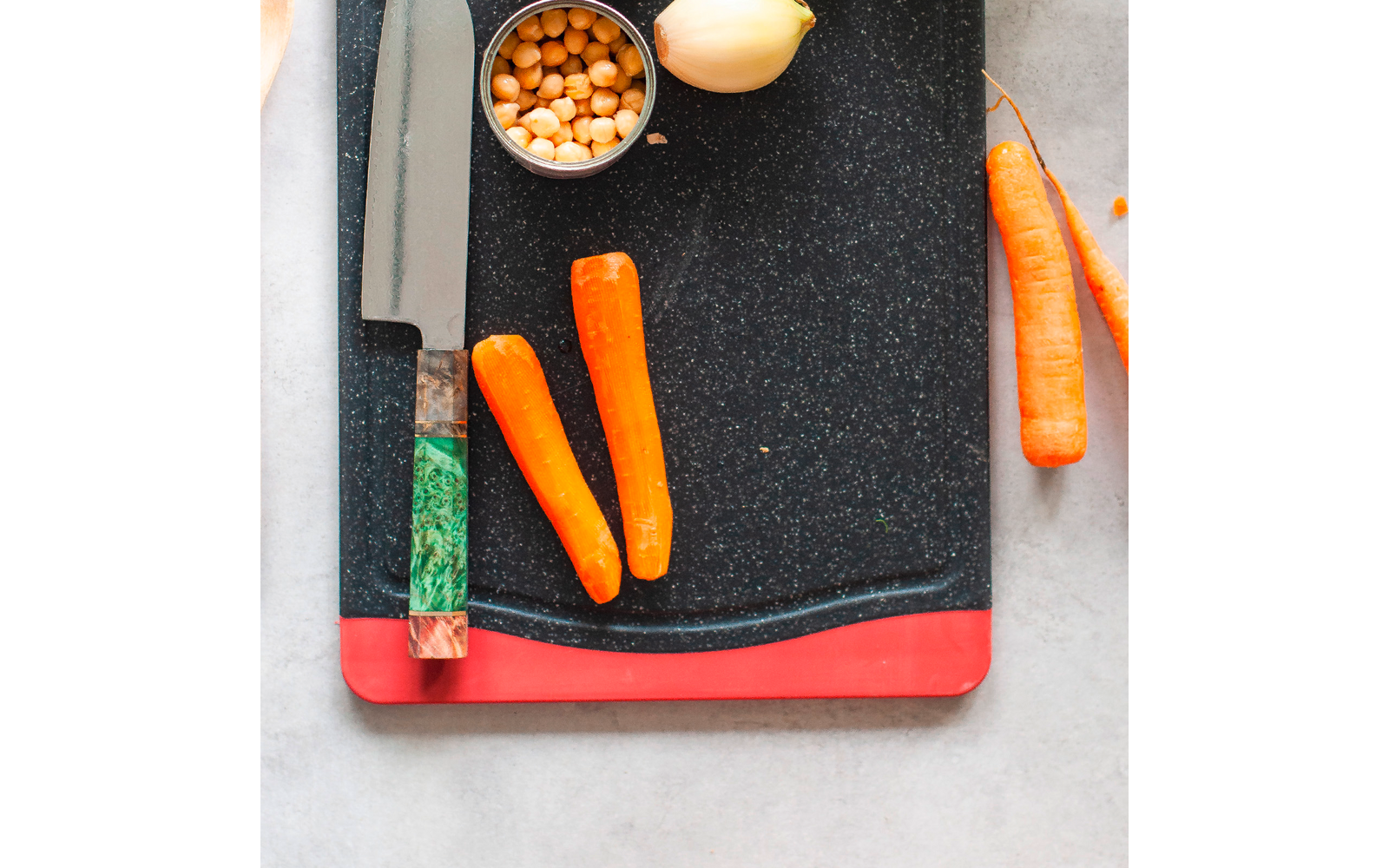 Schwarzes Schneidebrett mit rotem Rand, Messer mit grünem Griff, zwei geschälte Möhren auf dem Brett, Dose mit Kichererbsen und halbierte Zwiebel oben, ganze Möhre rechts
