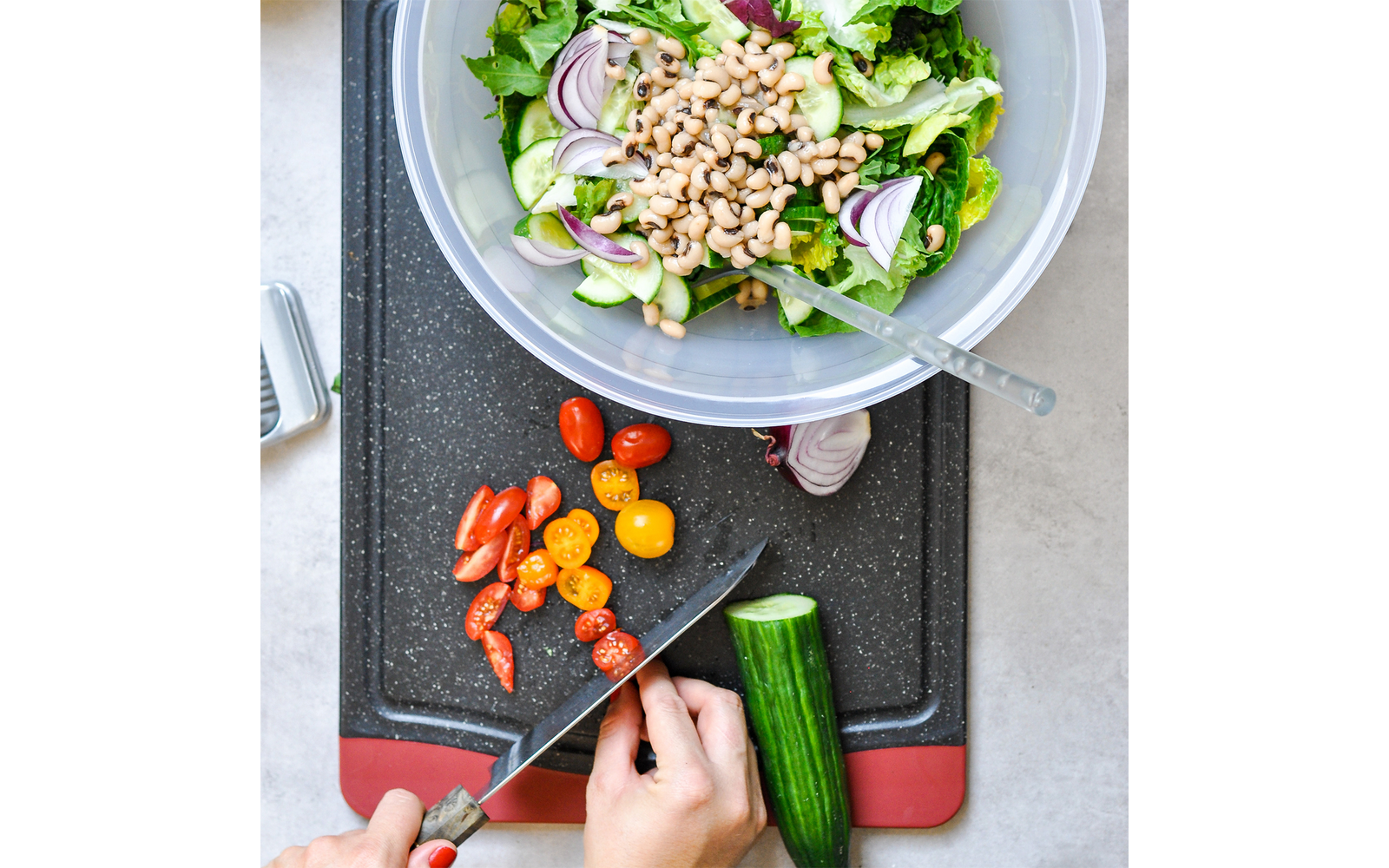 Vogelperspektive: Hände schneiden eine Gurke auf einem Schneidebrett; daneben geschnittene rote und gelbe Cherrytomaten. Oben eine Plastikschüssel mit gemischtem Salat, Gurkenscheiben, roten Zwiebelringen und hellen Bohnen