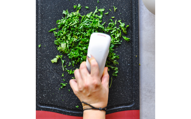 Hand mit Armband hält weißes Handgerät über gehackten grünen Kräutern auf schwarzem Schneidebrett