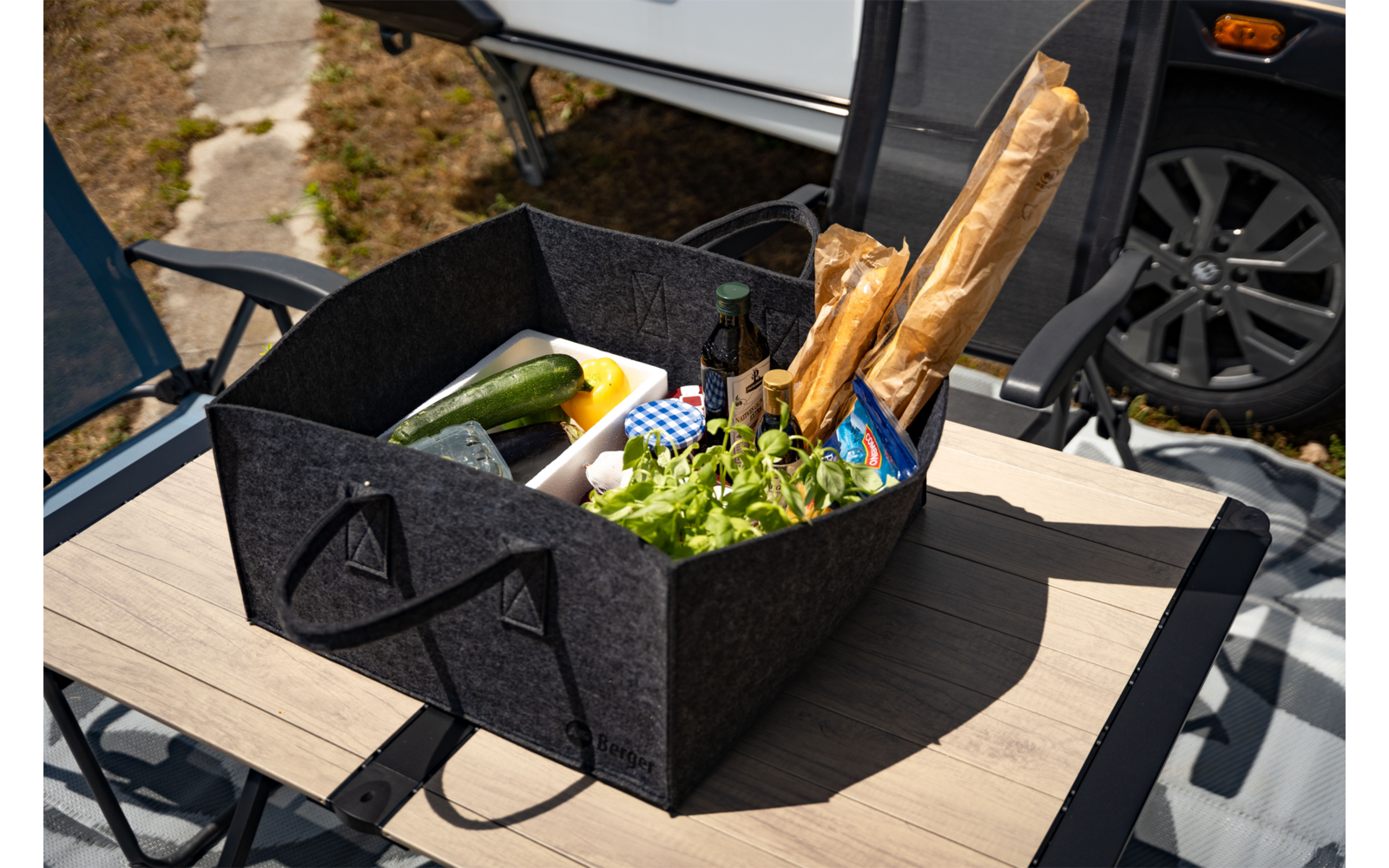 Schwarze Filztasche auf einem Holztisch mit zwei Baguettes in Papier, Kopfsalat, zwei Zucchini, gelber Butterdose, Flasche und blau-weiß kariertem Tuch; im Hintergrund Wohnwagen und Autoreifen