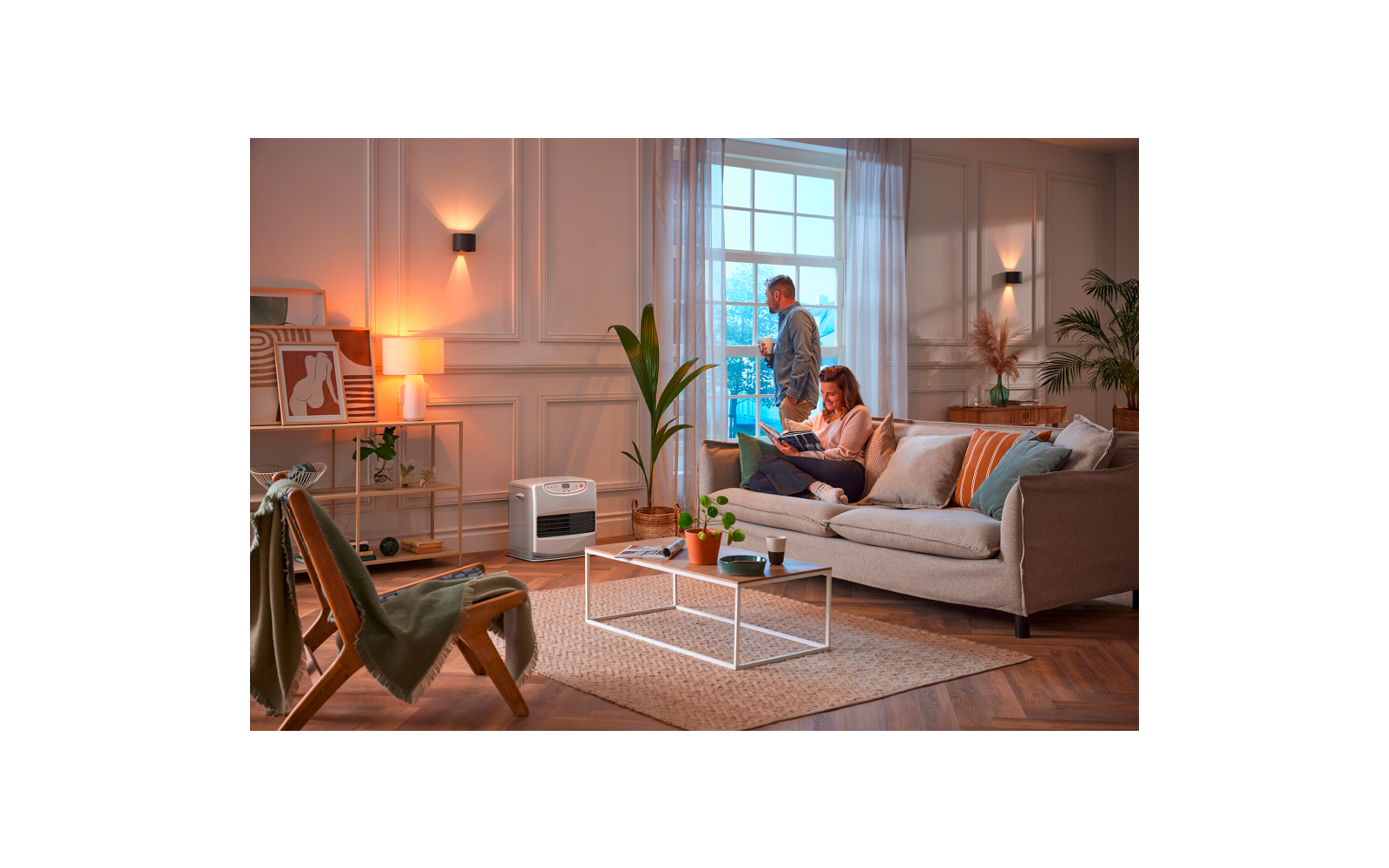 Two people in a living room: one sits on a sofa reading a book, the other stands at the window. Sofa with cushions, coffee table with cups, plants, shelving, wall lamps, rug and a white appliance