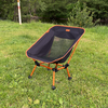 Une chaise avec armature orange et assise en filet noir sur l'herbe ; à l'arrière-plan des arbres et un plan d'eau ; à gauche un poteau blanc avec une marque orange