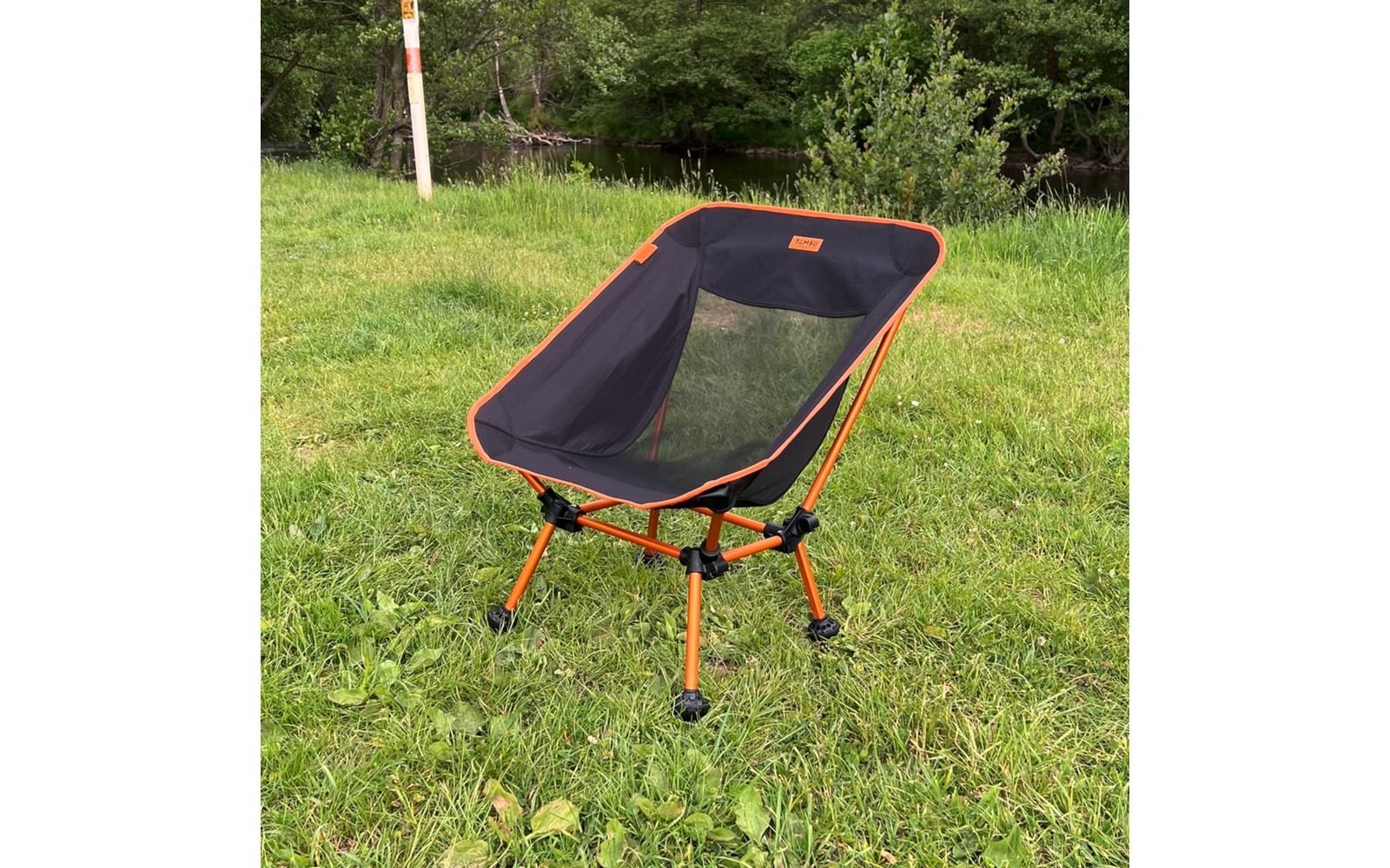 Une chaise avec armature orange et assise en filet noir sur l'herbe ; à l'arrière-plan des arbres et un plan d'eau ; à gauche un poteau blanc avec une marque orange