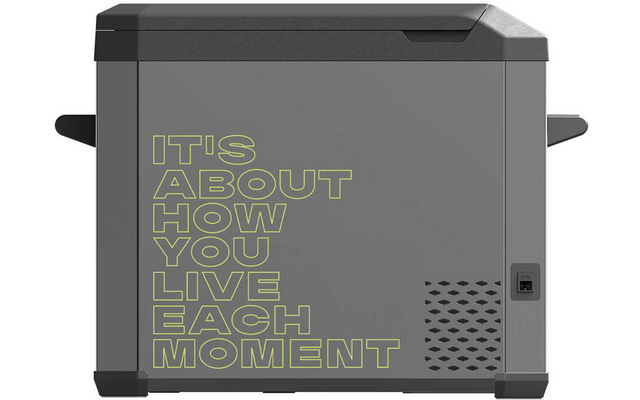 Graue rechteckige Box mit aufgedrucktem Text "IT'S ABOUT HOW YOU LIVE EACH MOMENT" in gelbgrüner Konturschrift, seitliche Griffe, Lüftungslöcher und kleiner rechteckiger Anschluss rechts