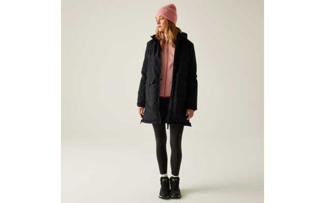 Frau trägt rosa Mütze, schwarzer Mantel, rosa Pullover, schwarze Hose und schwarze Schuhe vor hellem Hintergrund
