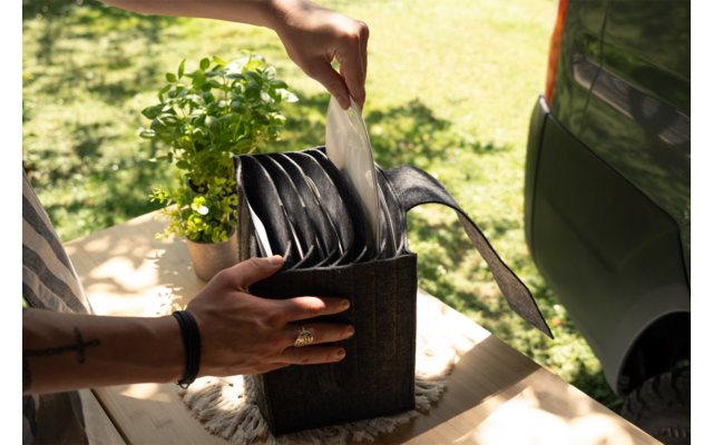 Hand trekt een doek uit een zwarte dispenser op een houten tafel; bloempot met plant links, groene auto en gras op de achtergrond