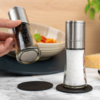 Hand houdt een gekanteld glas met hele zwarte peperkorrels en metalen deksel; daarnaast staat een vergelijkbaar glas met grof zout op zwarte onderzetters op een marmeren oppervlak; op de achtergrond een kom met eten, een stuk kaas en een plant