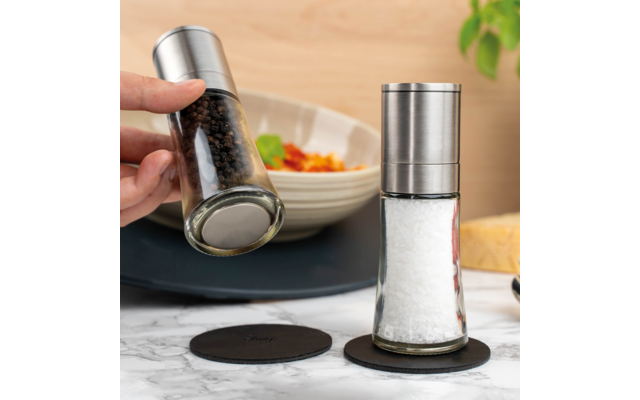 Hand houdt een gekanteld glas met hele zwarte peperkorrels en metalen deksel; daarnaast staat een vergelijkbaar glas met grof zout op zwarte onderzetters op een marmeren oppervlak; op de achtergrond een kom met eten, een stuk kaas en een plant