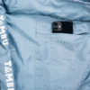 Light blue jacket lining with an inner pocket holding a black smartphone; white text 'TAMBU' on the hem