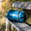 Blue compressed sleeping bag in a stuff sack with white text "TAMBU connect to nature" on a weathered wooden bench outdoors
