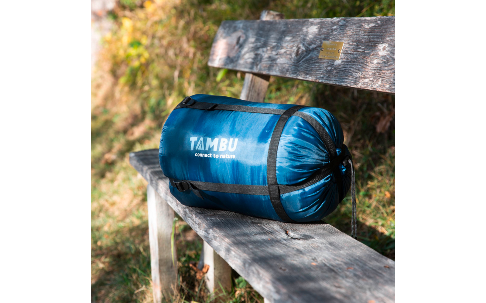 Blue compressed sleeping bag in a stuff sack with white text "TAMBU connect to nature" on a weathered wooden bench outdoors