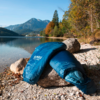 Blue sleeping bag on pebbles at the shore of a mountain lake, trees with autumn colors, mountains and clear sky in the background