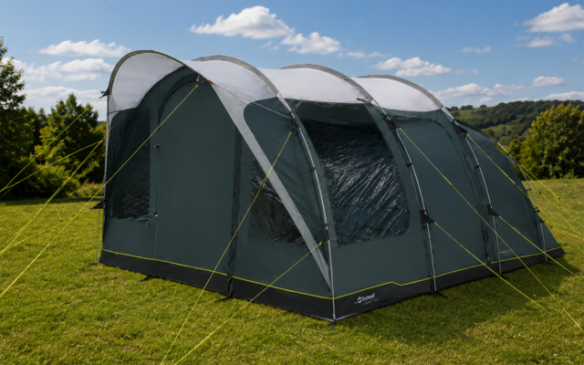 Grote grijs-groene tent met drie gebogen stokken, doorzichtige ramen en gele scheerlijnen op een grasveld, bomen en heuvels op de achtergrond onder een blauwe lucht met wolken