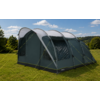 Grote grijs-groene tent met drie gebogen stokken, doorzichtige ramen en gele scheerlijnen op een grasveld, bomen en heuvels op de achtergrond onder een blauwe lucht met wolken