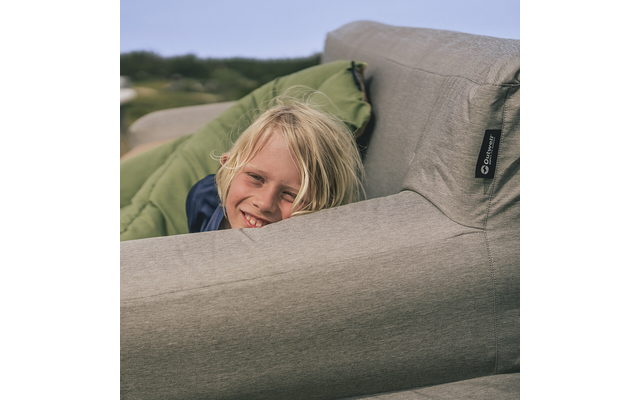 Enfant blond souriant regardant depuis un canapé gris, coussin vert derrière, paysage flou en arrière-plan