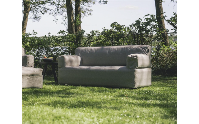 Canapé gris deux places sur l'herbe, à gauche un fauteuil partiellement visible et une petite table d'appoint, arbres et arbustes en arrière-plan