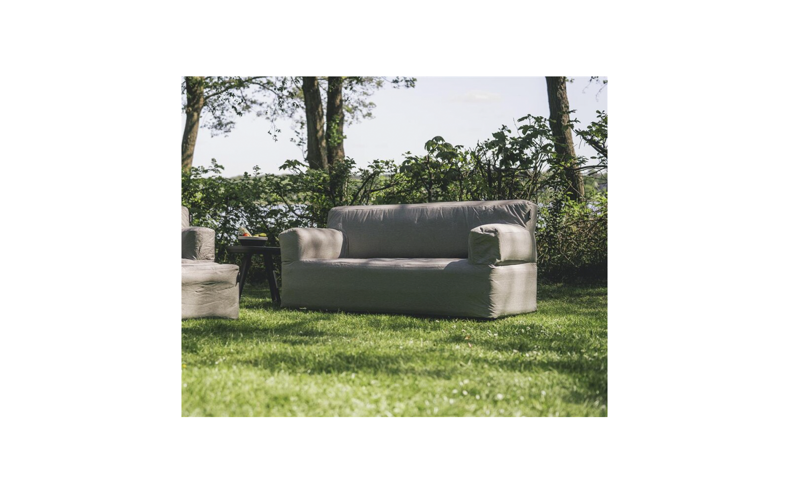 Canapé gris deux places sur l'herbe, à gauche un fauteuil partiellement visible et une petite table d'appoint, arbres et arbustes en arrière-plan
