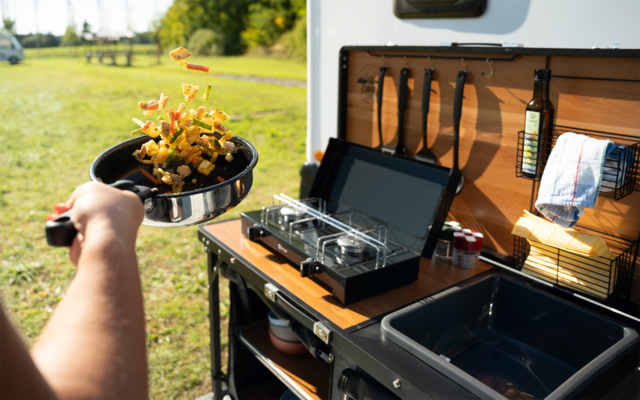 Arm hält Pfanne mit Gemüse über Campingkocher neben offener Wohnmobil-Küche, Wiese im Hintergrund