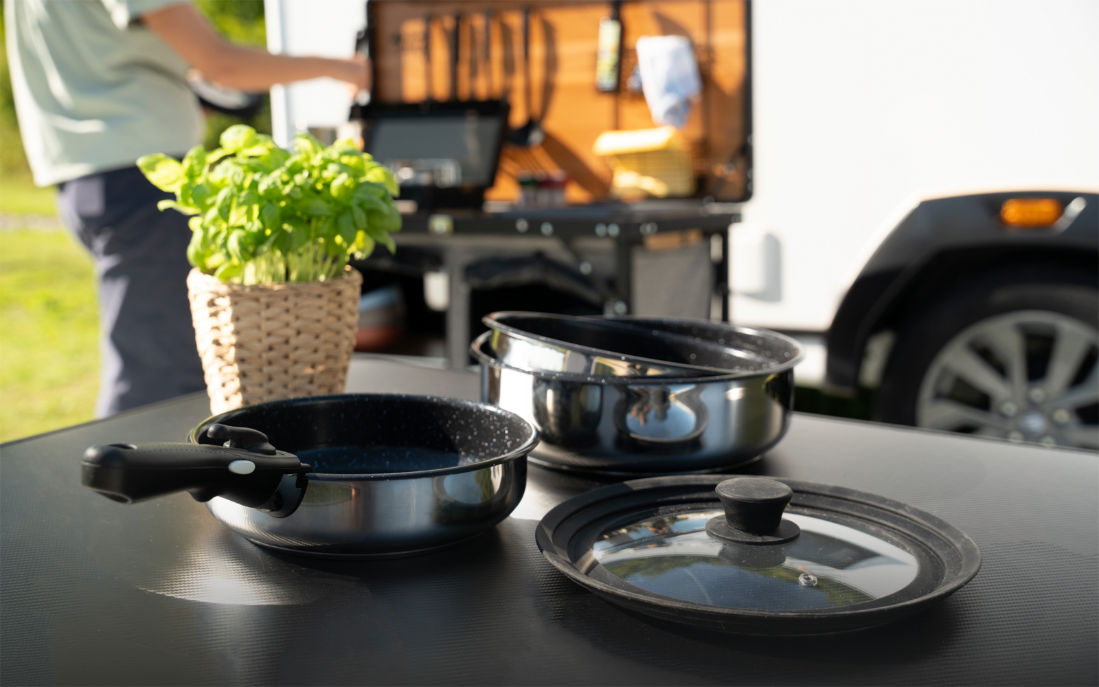 Drei Kochpfannen und ein Glasdeckel auf einem Tisch, neben einem Basilikum in einem geflochtenen Topf; im Hintergrund eine Person neben einem offenen Fahrzeugfach mit Campingkocher