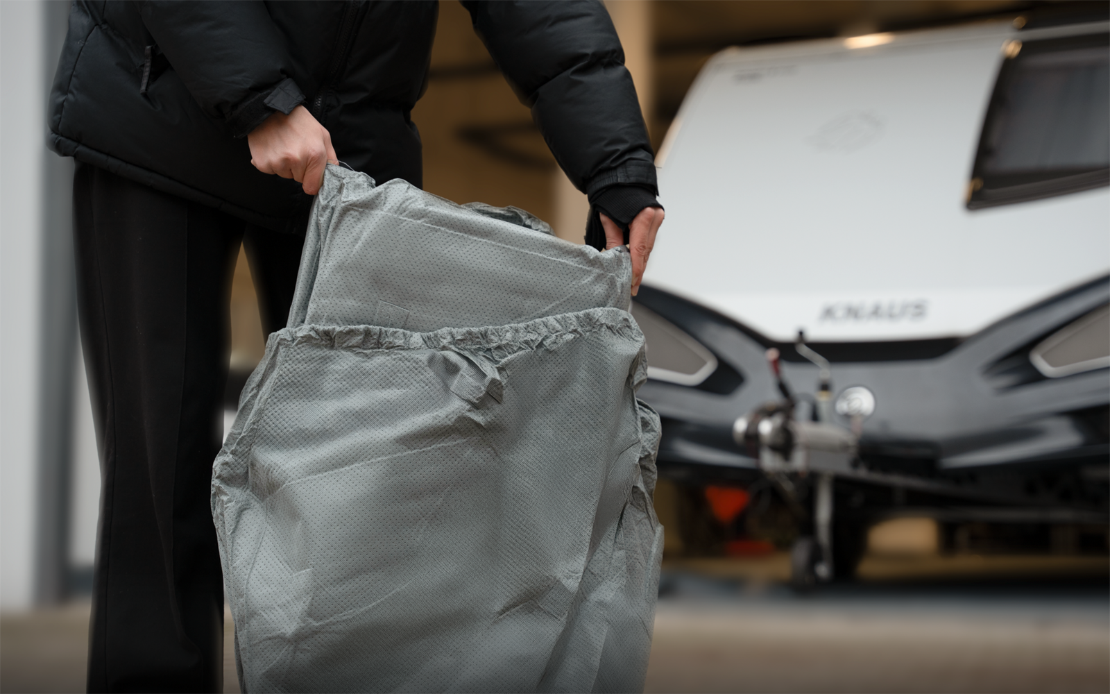 Hands of a person wearing a black jacket holding a folded gray fabric cover in front of a caravan