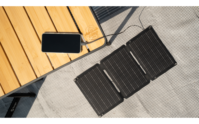 Ein faltbares Solarpanel liegt auf einem grauen Boden neben einem Holztisch mit einem Kabel und einem Stecker