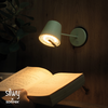 Wandlampe an Holzverkleidung leuchtet auf ein aufgeschlagenes Buch; eine Hand hält das Buch, Pflanzblätter links, Markenlogo unten links
