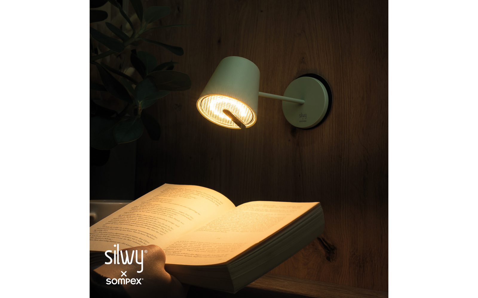 Wandlampe an Holzverkleidung leuchtet auf ein aufgeschlagenes Buch; eine Hand hält das Buch, Pflanzblätter links, Markenlogo unten links