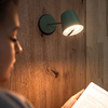 Linkes Profil einer Person, Hand hält ein aufgeschlagenes Buch; an Holzwand montierte Lampe leuchtet