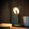Grüne Schreibtischlampe eingeschaltet, links ein offenes Buch, rechts eine weiße Tasse, Holzoberfläche und Holzwand im Hintergrund