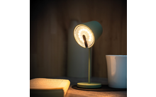 Lampe de bureau verte allumée, livre ouvert à gauche, tasse blanche à droite, surface en bois et mur en bois en arrière-plan