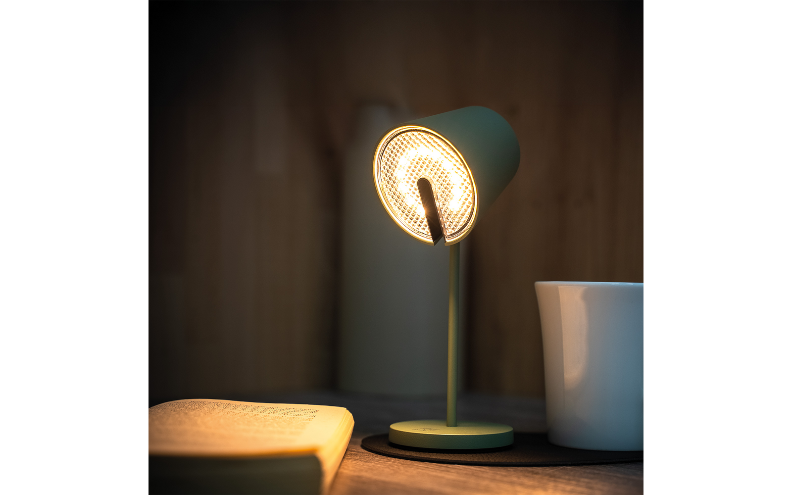 Grüne Schreibtischlampe eingeschaltet, links ein offenes Buch, rechts eine weiße Tasse, Holzoberfläche und Holzwand im Hintergrund