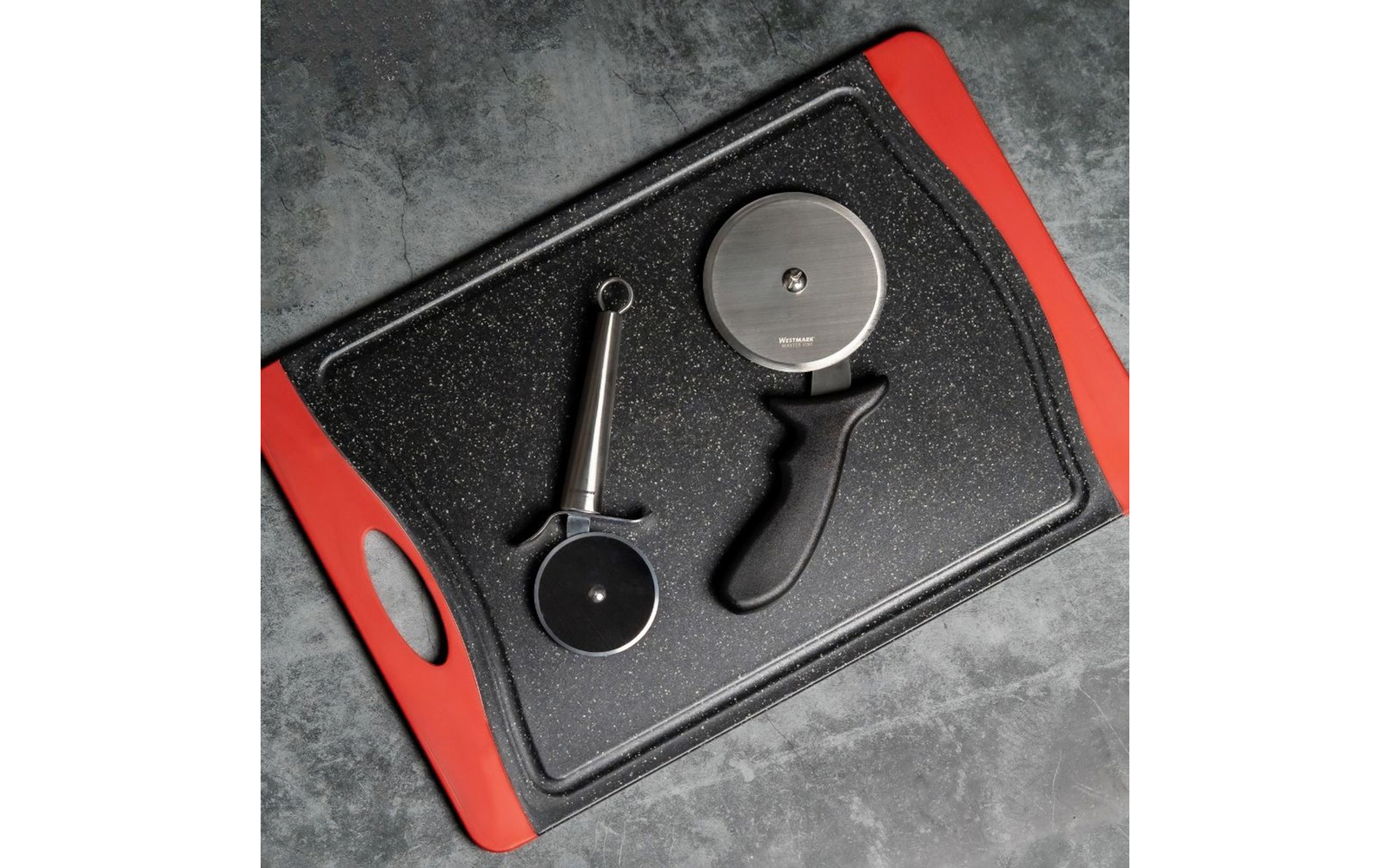Rechteckiges schwarzes Schneidebrett mit roten Griffen auf grauem Untergrund, darauf zwei Pizzaschneider: einer mit Metallgriff und kleiner runder Klinge, einer mit schwarzem Griff und großer runder Metallklinge