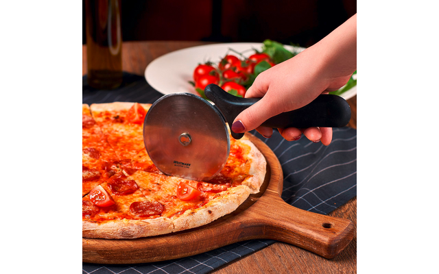 Main avec vernis sombre tenant une roulette à pizza sur une pizza garnie de tranches de tomate et de pepperoni sur une planche en bois ; assiette de tomates cerises et bouteille d'huile en arrière-plan