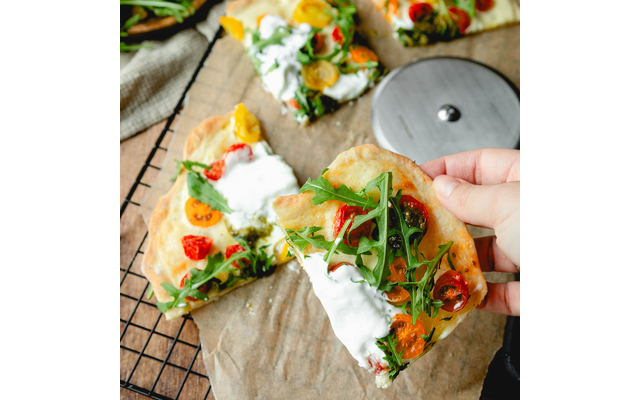 Main tenant un morceau de pain plat avec garniture blanche crémeuse, roquette et tomates cerises jaunes et rouges coupées en deux ; d'autres morceaux et une roulette à pizza ronde sur une grille de refroidissement