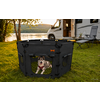 Dog lying inside a black foldable pet playpen on grass; background shows a lake, trees, and a white motorhome with an open door and two camping chairs