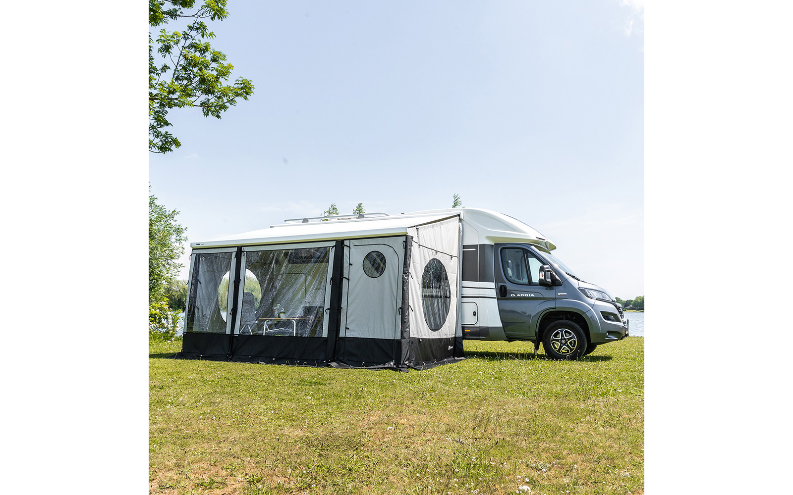Weißes Wohnmobil mit angebautem grauem Vorzelt auf einer Wiese neben einem Gewässer; rundes Fenster im Vorzelt und Stühle sichtbar im Inneren