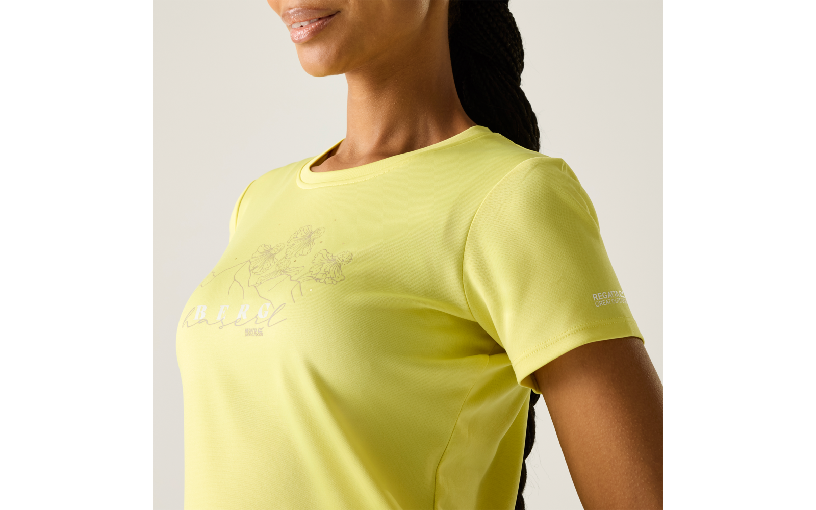 Vue partielle du torse d'une personne portant un t-shirt jaune à manches courtes, avec un imprimé floral sur la poitrine et un petit logo sur la manche, cheveux tressés attachés vers l'arrière