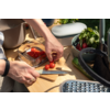 Zwei Hände schneiden Kirschtomaten mit einem Messer auf einem Holzbrett; daneben ein Glasbehälter mit Tomaten, eine Schüssel mit Radieschen und Kräutern, eine Reibe und eine Pflanze