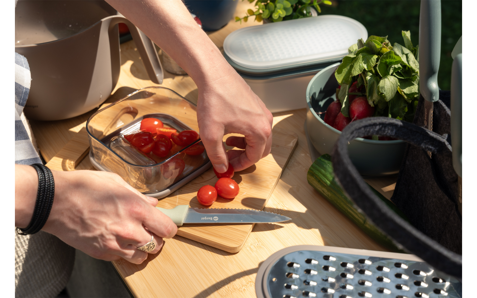 Zwei Hände schneiden Kirschtomaten mit einem Messer auf einem Holzbrett; daneben ein Glasbehälter mit Tomaten, eine Schüssel mit Radieschen und Kräutern, eine Reibe und eine Pflanze