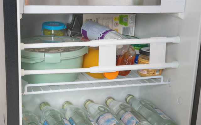 Innenraum eines Kühlschranks mit einer grünen Kunststoffdose, einem umgekippten Joghurtbecher, einer Orange, Gläsern und mehreren liegenden Glasflaschen