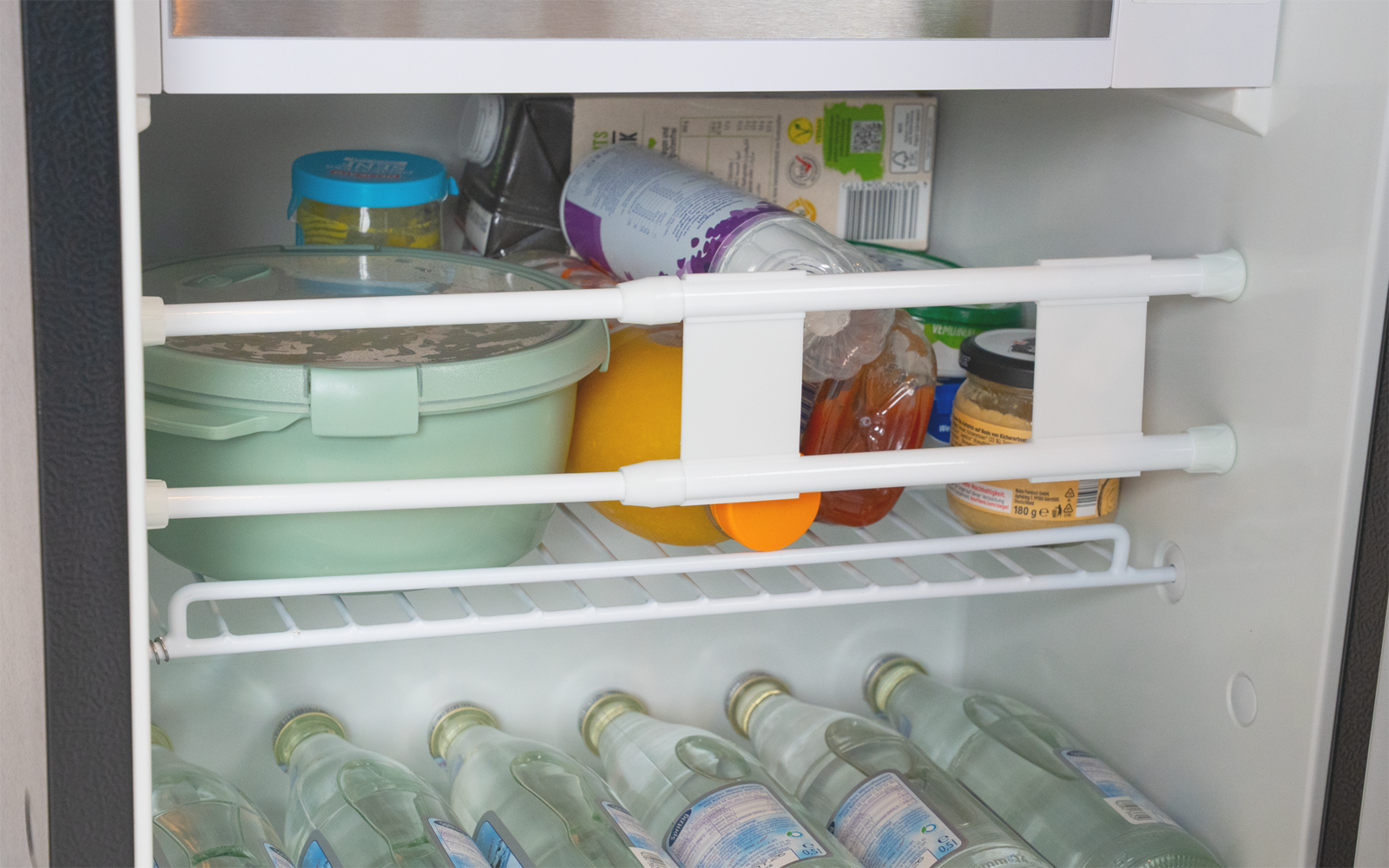 Innenraum eines Kühlschranks mit einer grünen Kunststoffdose, einem umgekippten Joghurtbecher, einer Orange, Gläsern und mehreren liegenden Glasflaschen