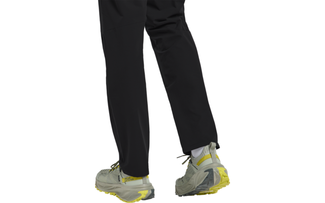 Beine einer Person in schwarzen Hosen und grauen Sportschuhen mit gelben Akzenten auf weißem Hintergrund