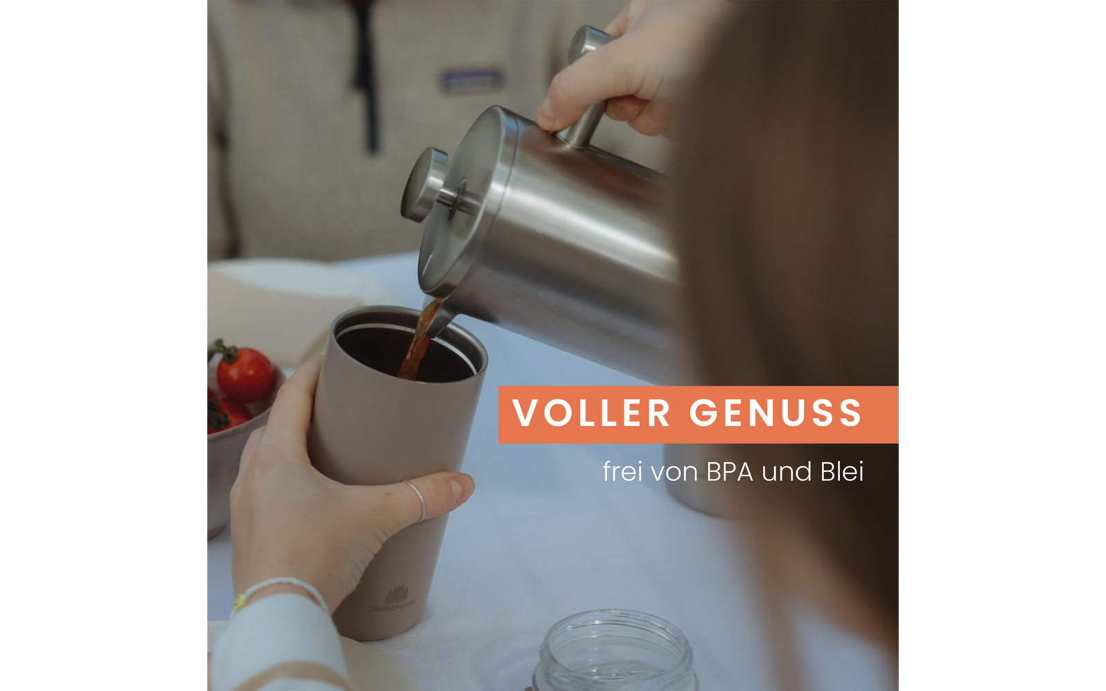 Hände gießen eine dunkle Flüssigkeit aus einer Edelstahlkanne in einen Thermobecher; im Hintergrund Glas, eine Schale mit einer Tomate und eine unscharfe Person; Schriftzug 'VOLLER GENUSS' und 'frei von BPA und Blei'