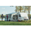 Twee personen naast een caravan met voortent; één persoon draagt een kind op de schouders. Caravan op gras bij een meer, bomen op de achtergrond, tassen op de grond en een fiets met fietsendrager zichtbaar