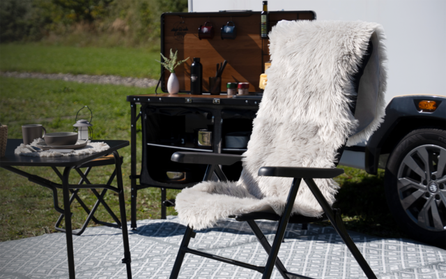 Faltstuhl mit weißem, flauschigem Überwurf auf graumusterter Matte; daneben runder Klapptisch mit Teller, Tasse und Laterne; offener Holz-Kasten auf schwarzem Gestell an einem Fahrzeug, Fahrzeugrad und Rasen im Hintergrund