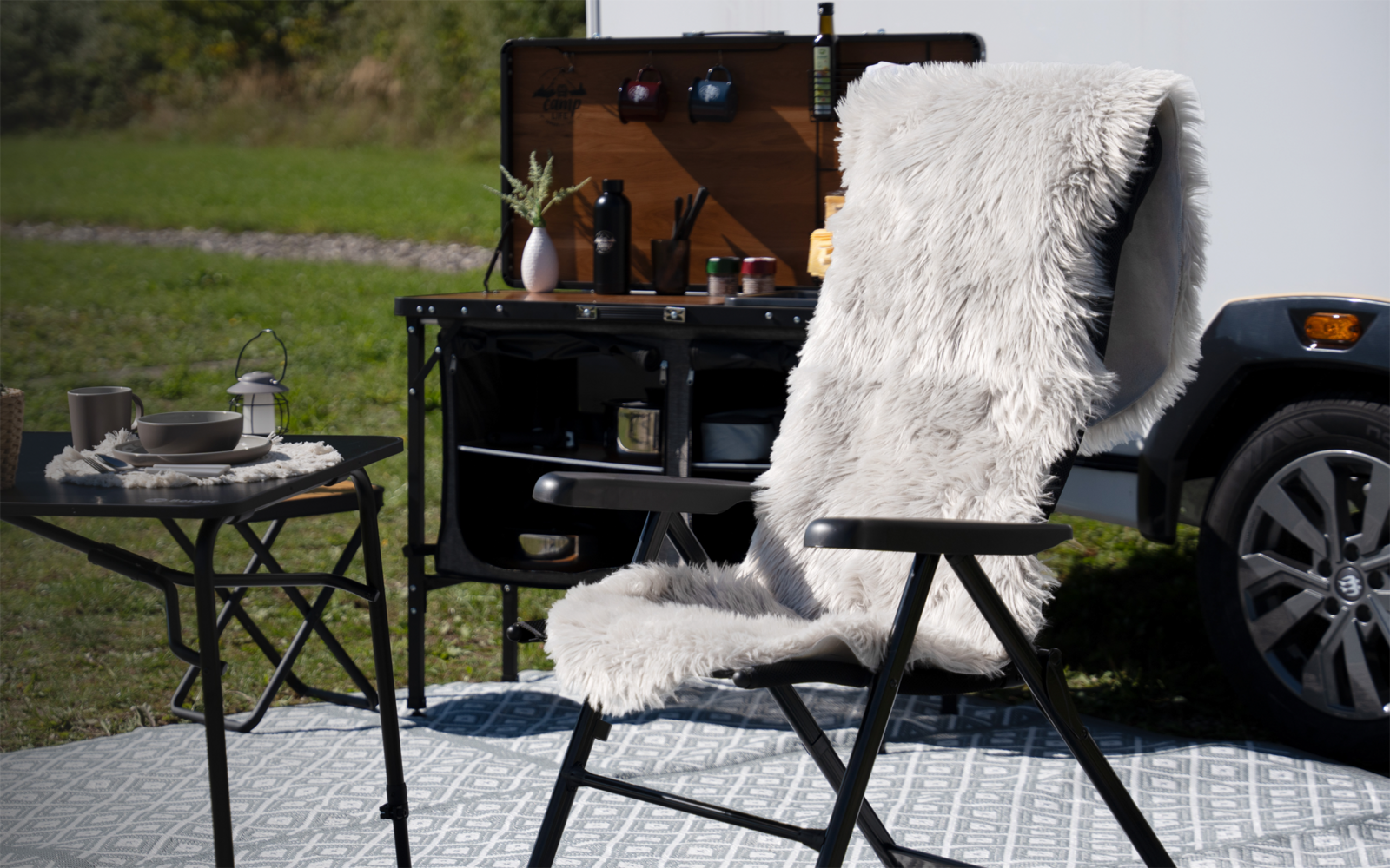 Faltstuhl mit weißem, flauschigem Überwurf auf graumusterter Matte; daneben runder Klapptisch mit Teller, Tasse und Laterne; offener Holz-Kasten auf schwarzem Gestell an einem Fahrzeug, Fahrzeugrad und Rasen im Hintergrund